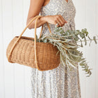 Woman in floral dress carrying a handwoven rattan market basket with dual arched handles and fresh eucalyptus