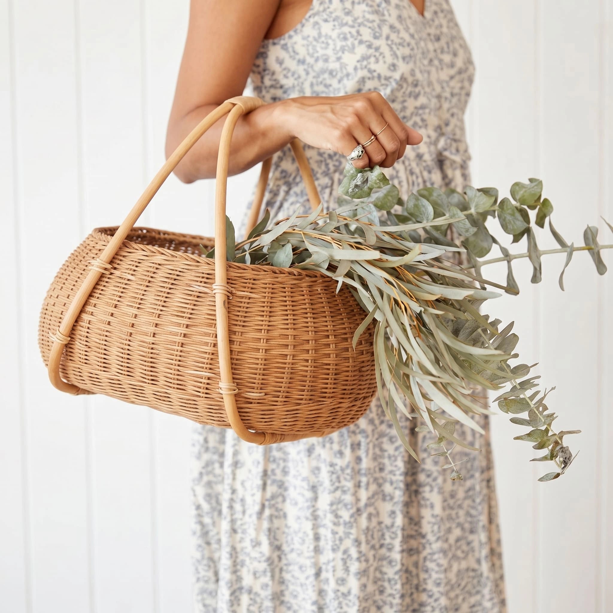 Woman in floral dress carrying a handwoven rattan market basket with dual arched handles and fresh eucalyptus